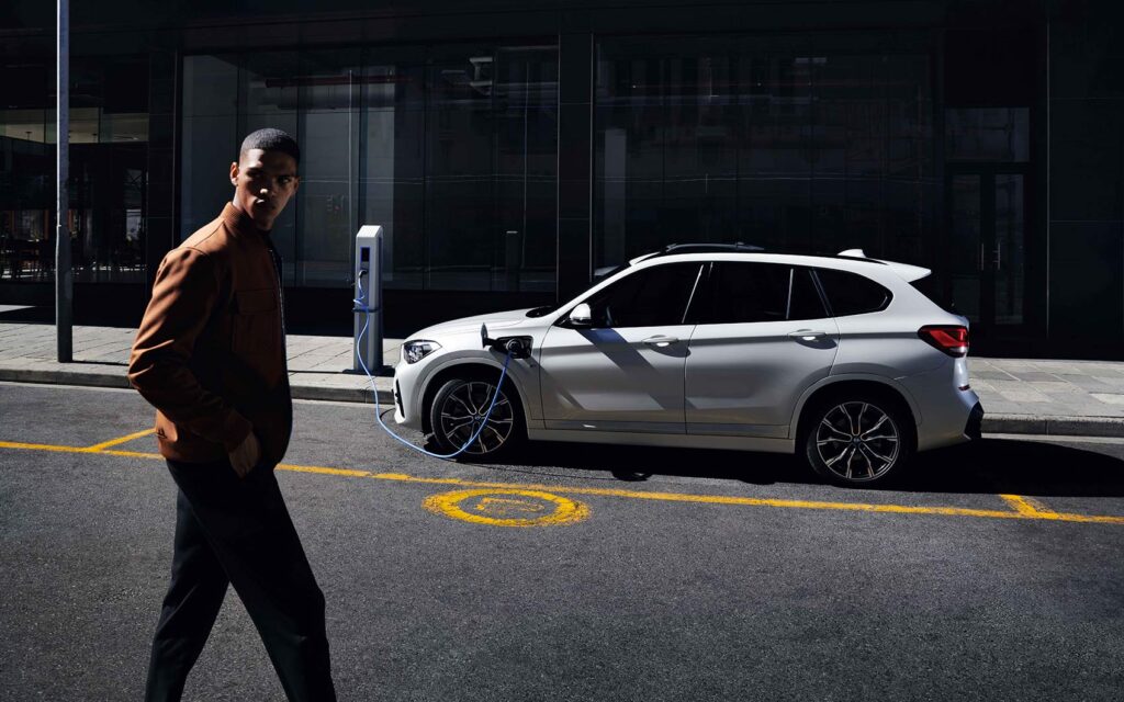 Weißer BMW steht am Straßenrand und wird per Ladekabel geladen. Im Vordergrund geht ein junger Mann mit brauner Jacke vorbei.